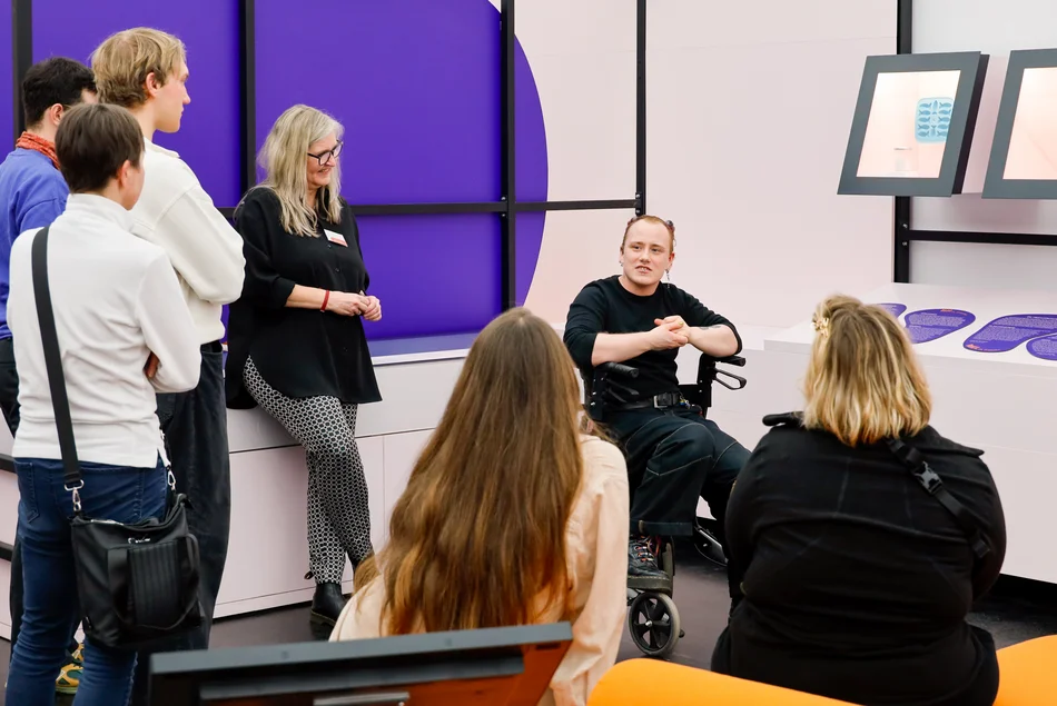Eine Person im Rollstuhl spricht mit einer Gruppe über eine Objekt im Studio "Bin ich schön!"