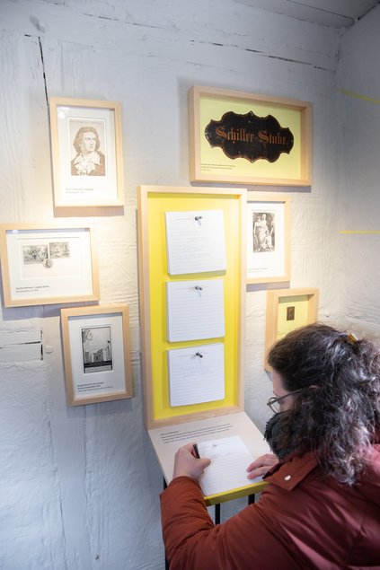 An einer weißen Ausstellungswand hängen mehrere kleine Bilder sowie ein längerer Bilderrahmen, an dessen unteres Ende sich eine kleine Schreibfläche angliedert. Eine Frau steht davor und schreibt etwas.