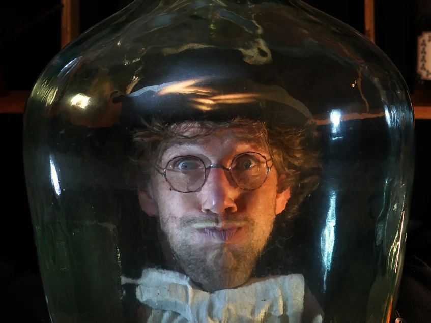 Das Bild zeigt eine Person mit einem Zylinder auf dem Kopf und einer Schleife am Hals in einem runden Glas. Die Person hält die Luft an.