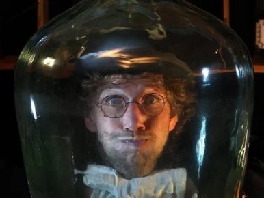 Das Bild zeigt eine Person mit einem Zylinder auf dem Kopf und einer Schleife am Hals in einem runden Glas. Die Person hält die Luft an.