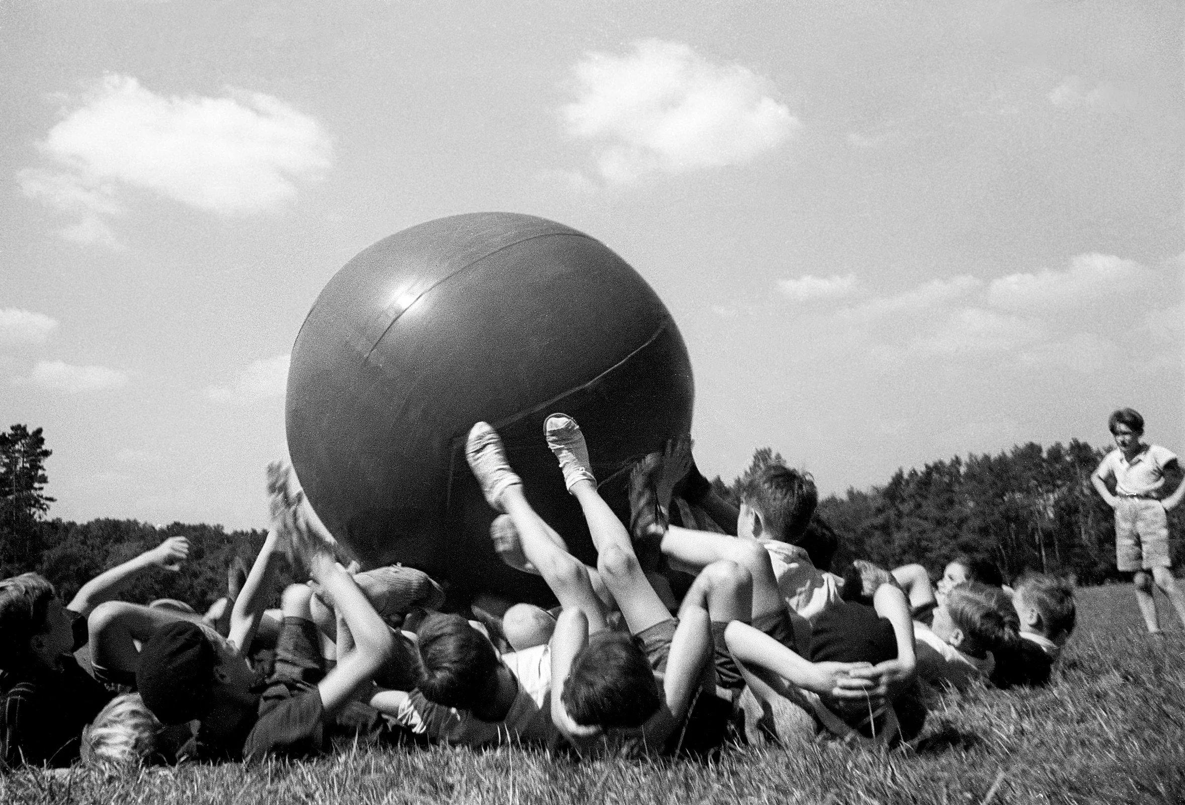 Fred Stein, „Big Ball“ Historische Schwarzweißfotografie von auf einer Wiese liegenden Kindern, die mit ihren Füßen mit einem riesigen Ball spielen