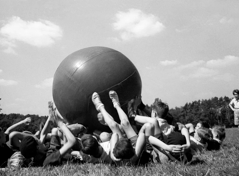 Historische Schwarzweißfotografie von auf einer Wiese liegenden Kindern, die mit ihren Füßen mit einem riesigen Ball spielen