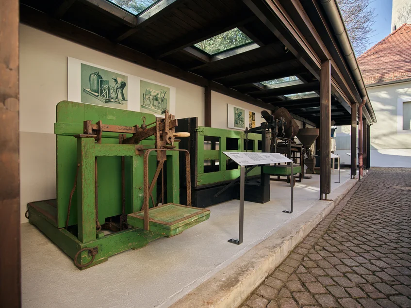 Das Foto zeigt die im Außenbereich des Museumskomplexes ausgestellten Großwaagen. An der Wand hinter den Waagen hängen jeweils Reproduktionen historischer grafischer Abbildungen der entsprechenden Waagen, davor ist ein Aufsteller mit Texten zu den Exponaten installiert. Zentral im Bild ist eine riesige, grün bemalte Getreidewaage, dahinter eine Viehwaage.