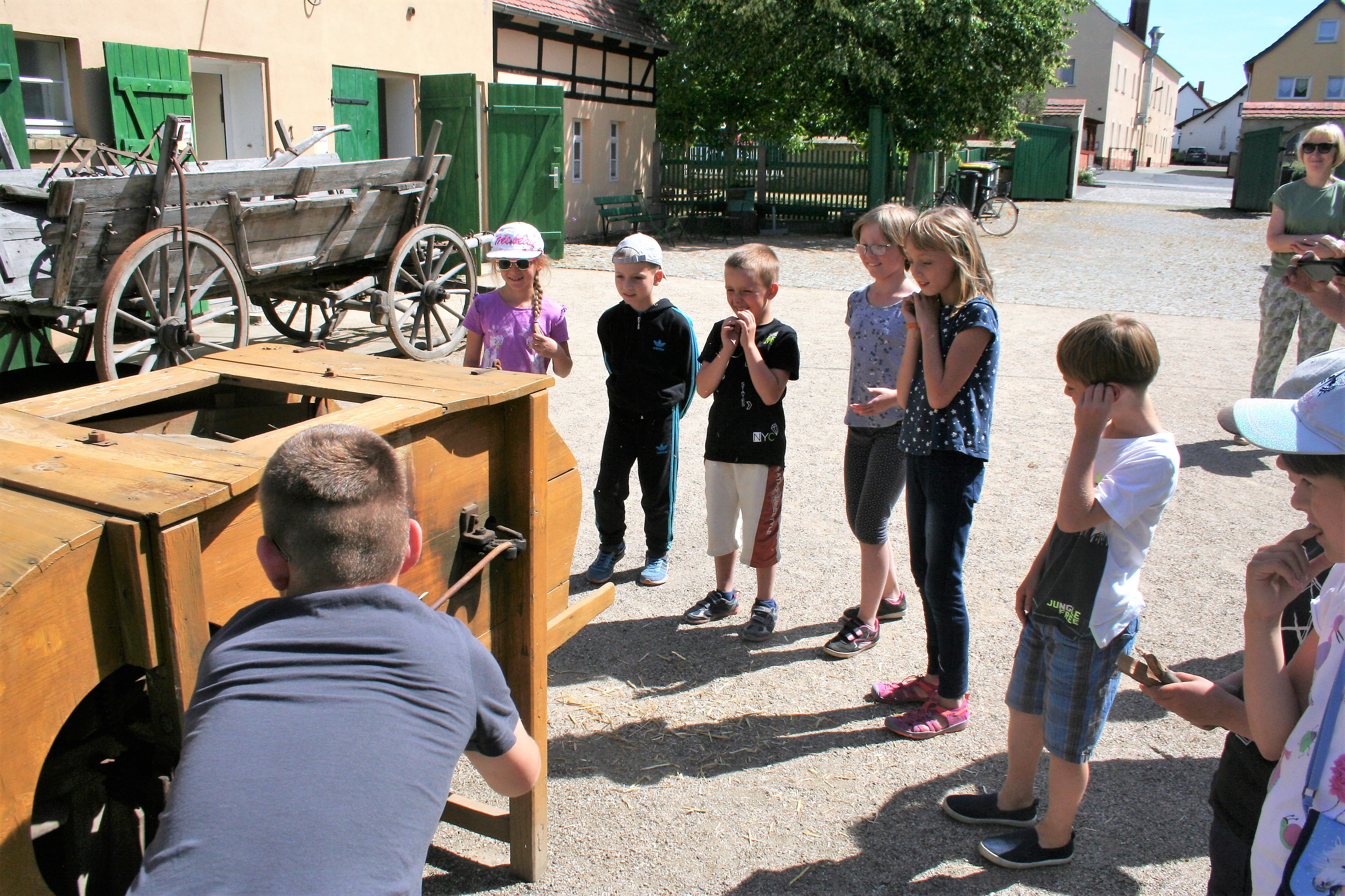 Eine Grundschulgruppe und ihre erwachsene Begleiterin stehen im Außenbereich des Museums, die Kinder inspizieren ein großes hölzernes Gerät namens Windfege.