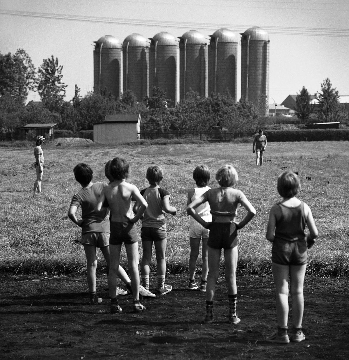 Schwarzweißfotografie einer sommerlichen Szene, in der mit dem Rücken zum Fotografen stehende, in Sportsachen gekleidete Kinder auf einer Wiese stehen und in Richtung zweier Erwachsener blicken, im Hintergrund stehen markant sechs Silos.