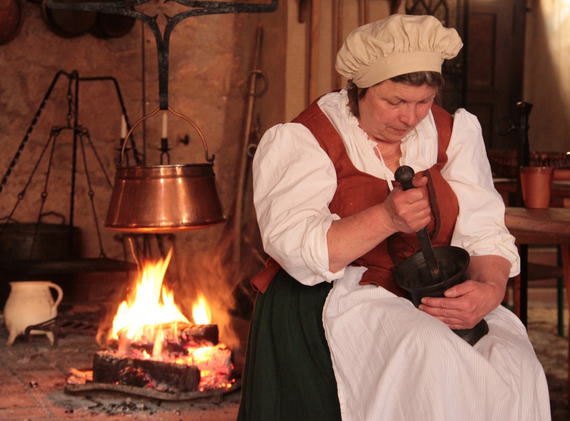 Eine im Kostüm einer historischen Hofköchin gekleidete Frau sitzt in der Schlossküche am Feuer und hält einen Mörser in den Händen.