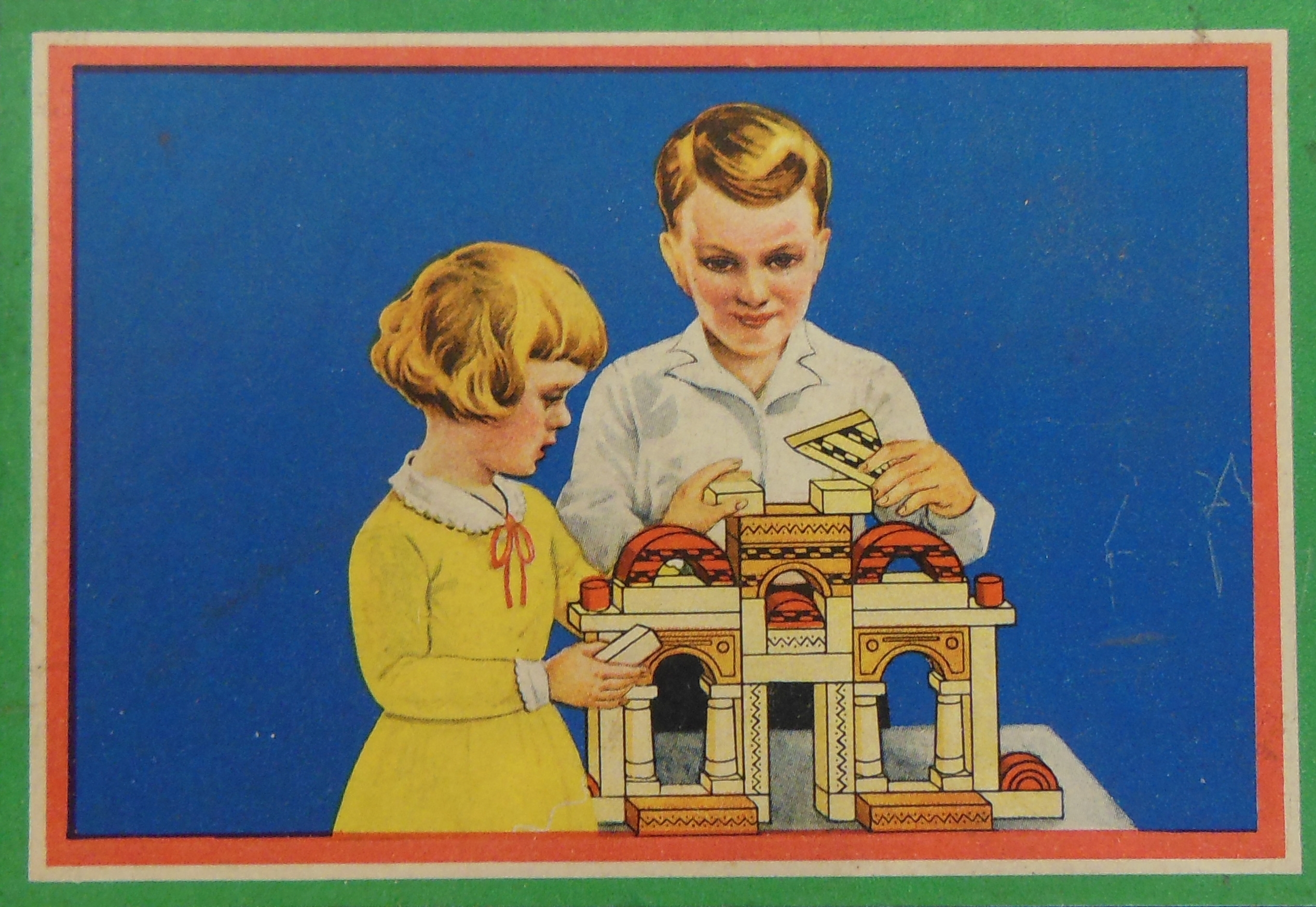 Foto des Deckels eines historischen Baukastens. Das Motiv zeigt ein Mädchen und einen Jungen, die gemeinsam Holzklötze zu einem Miniatur-Bauwerk fügen.