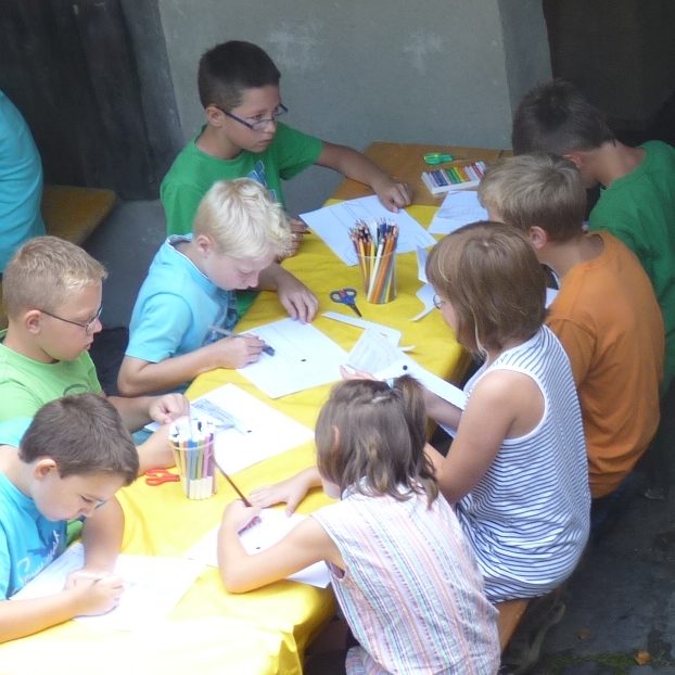 Kinder sitzen an einem Tisch und arbeiten mit Buntstiften, Schere und Leim auf Papier.