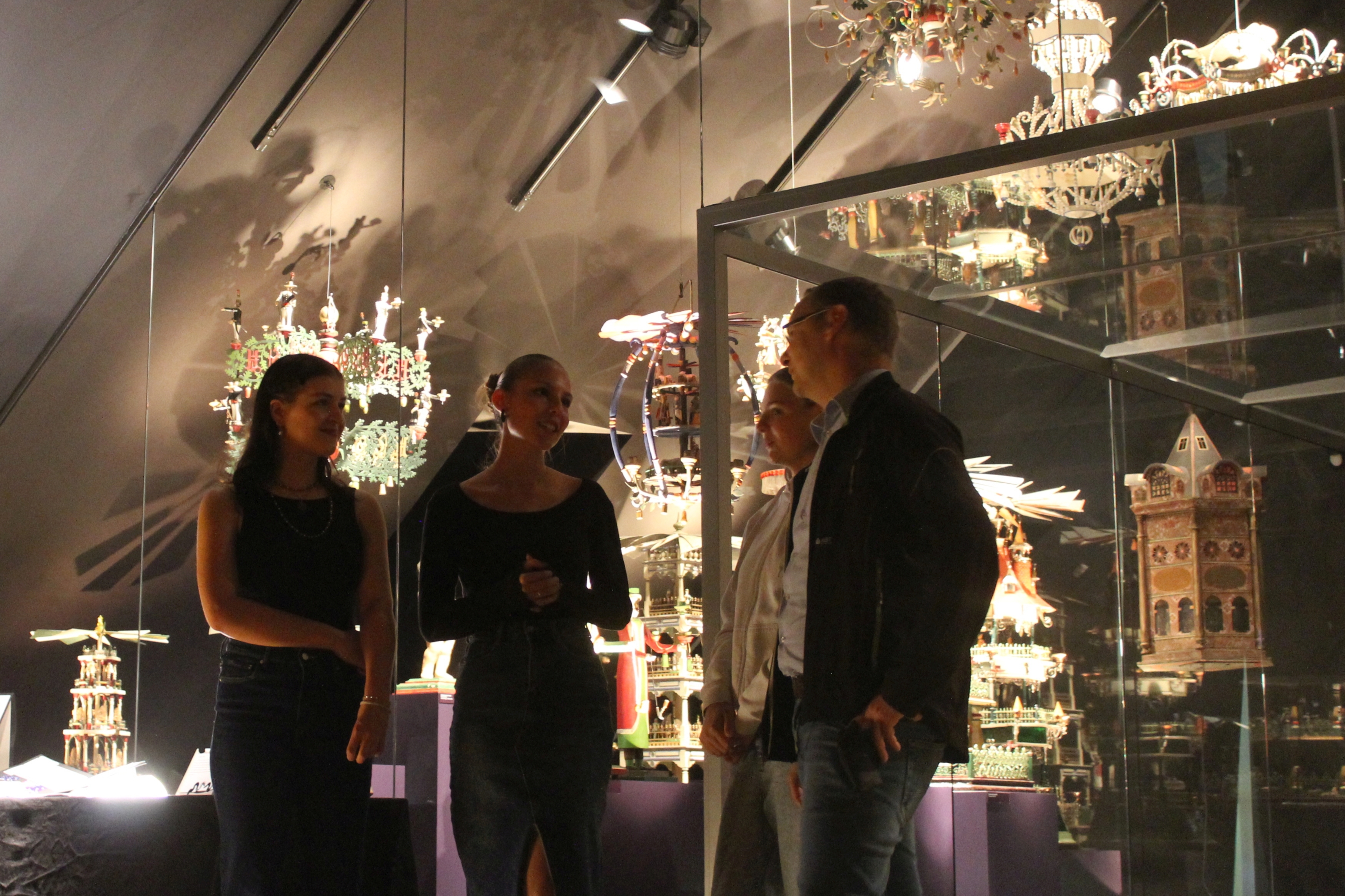 Vier Personen stehen in einem Ausstellungsraum, in dem eine Vielzahl erzgebirgischer Weihnachtspyramiden und andere Schnitzkunst präsentiert werden.