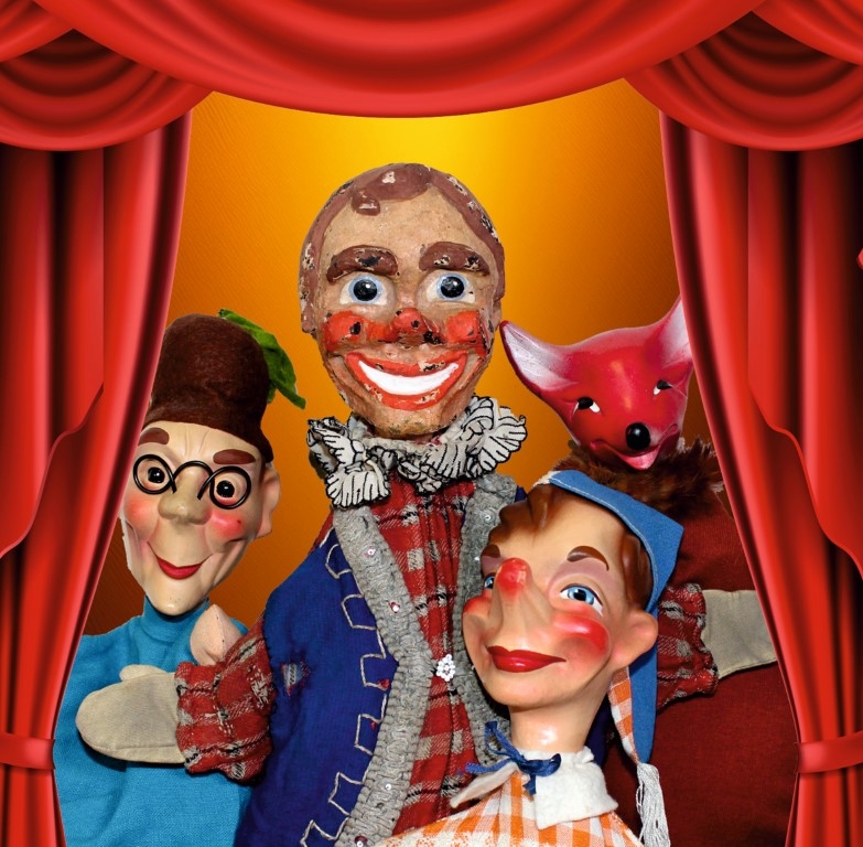 Auf einer Bühne mit geöffnetem roten Vorhang befinden sich vier Handpuppen – Kasper, Fuchs und zwei weitere Charaktere.