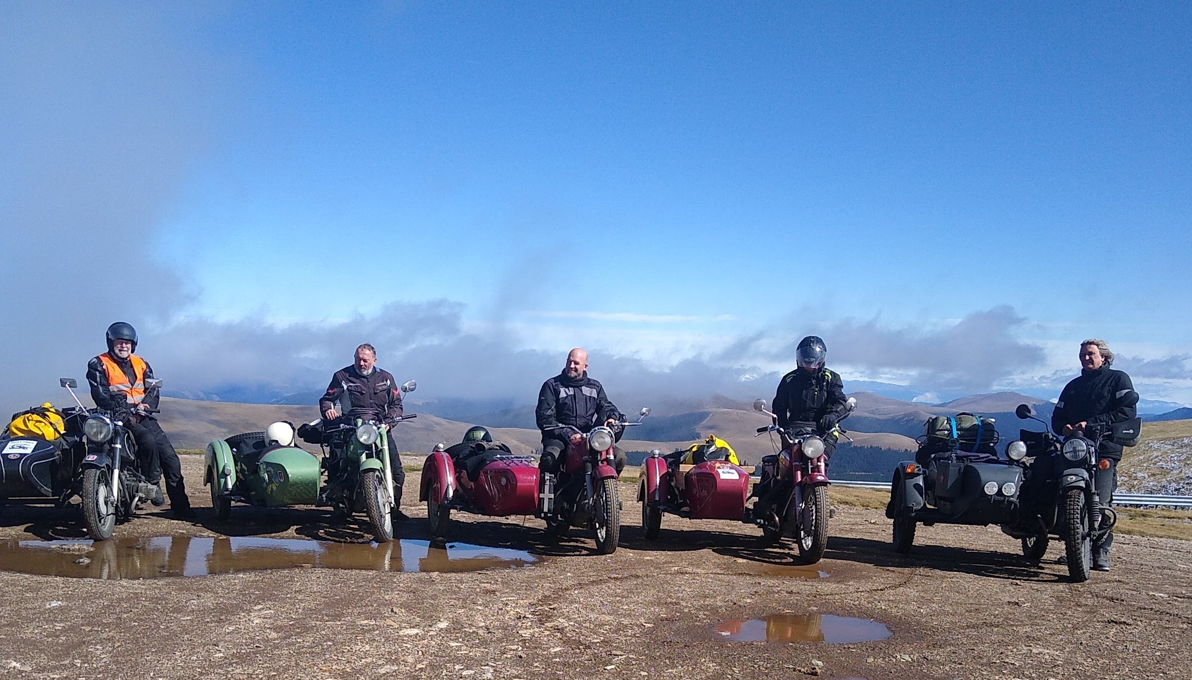 Fünf Motorradfahrer stehen nebeneinander mit ihren historischen Motorrädern mit Beiwagen in einer kargen Hochgebirgslandschaft.