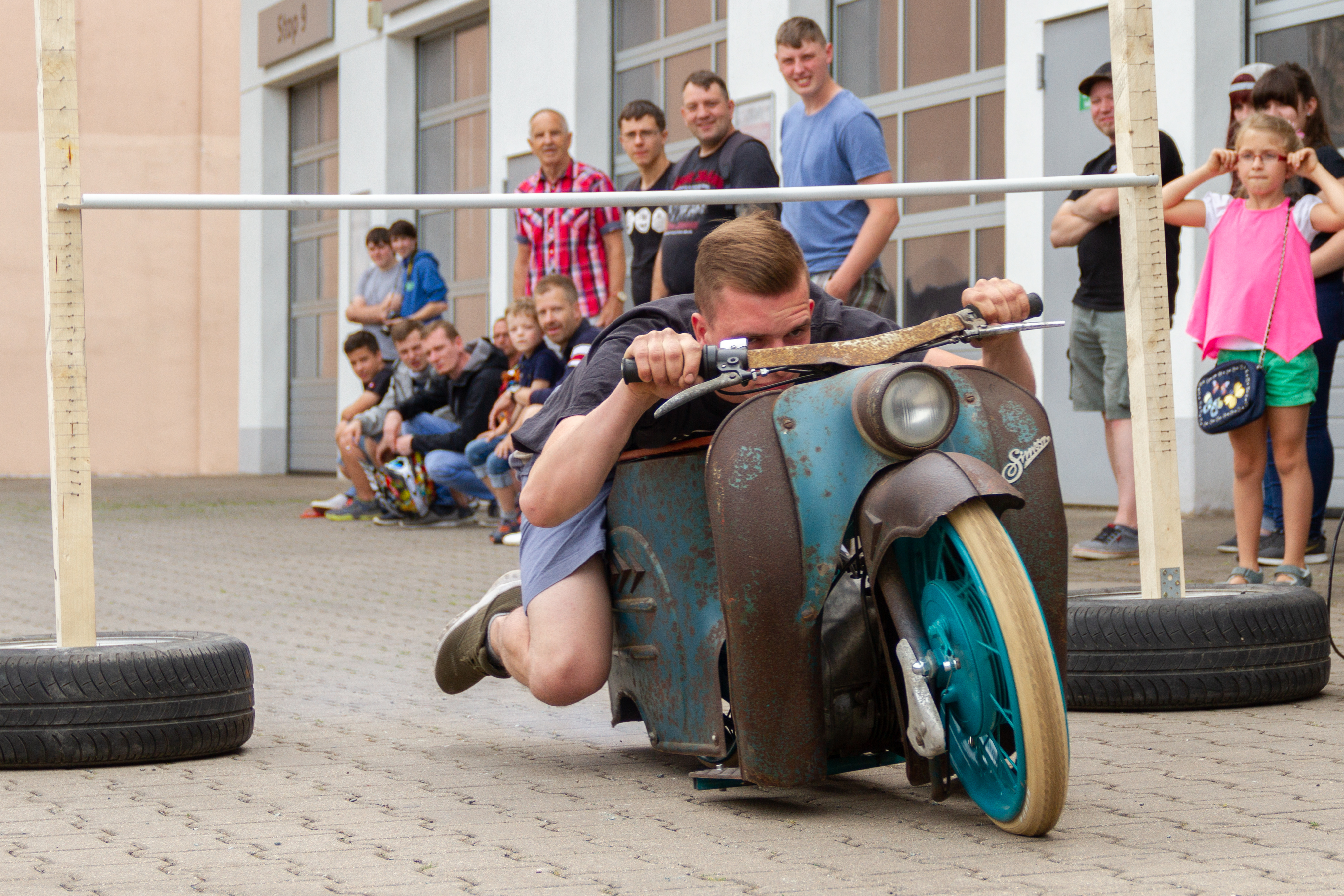 Foto eines jungen Mannes auf einem sehr alten Simson-Moped, der geduckt unter einer niedrig gehängten Stange hindurchfährt. Im Hintergrund stehen Menschen und schauen zu.