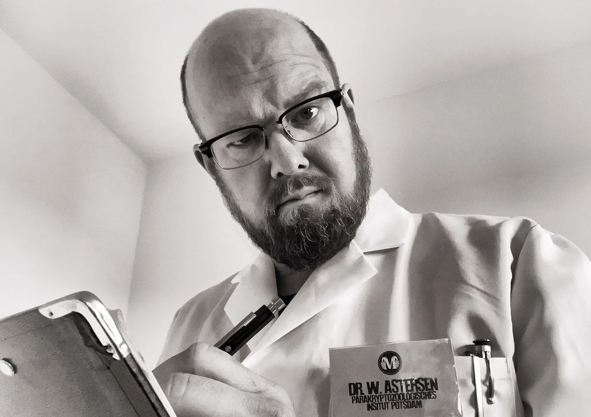 Ein Mann mittleren Alters mit Glatze, Bart, Brille, weißem Kittel und Klemmbrett blickt skeptisch in die Kamera.