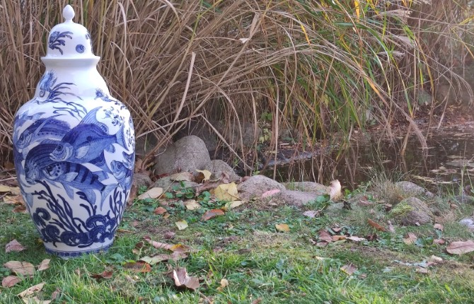 Weiße Vase mit Fisch- und Unterwassermotiven in Blautönen steht an der Uferböschung eines Gewässers