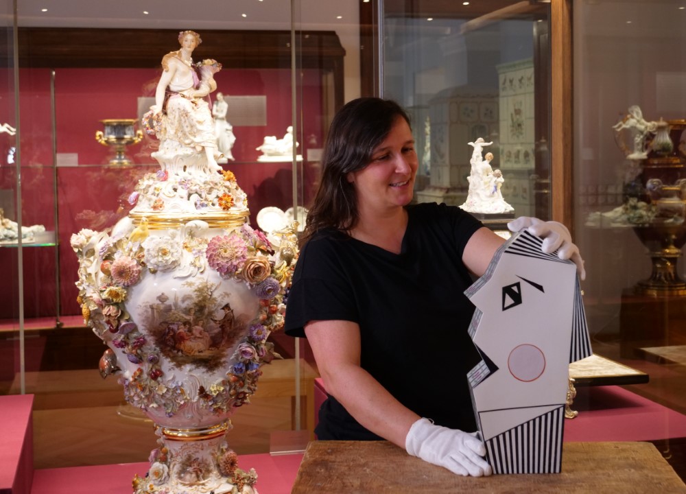 Eine Kuratorin mit weißen Handschuhen präsentiert eine moderne Büste aus Meissener Porzellan. Im Hintergrund steht eine riesige, reich verzierte historische Porzellanvase.