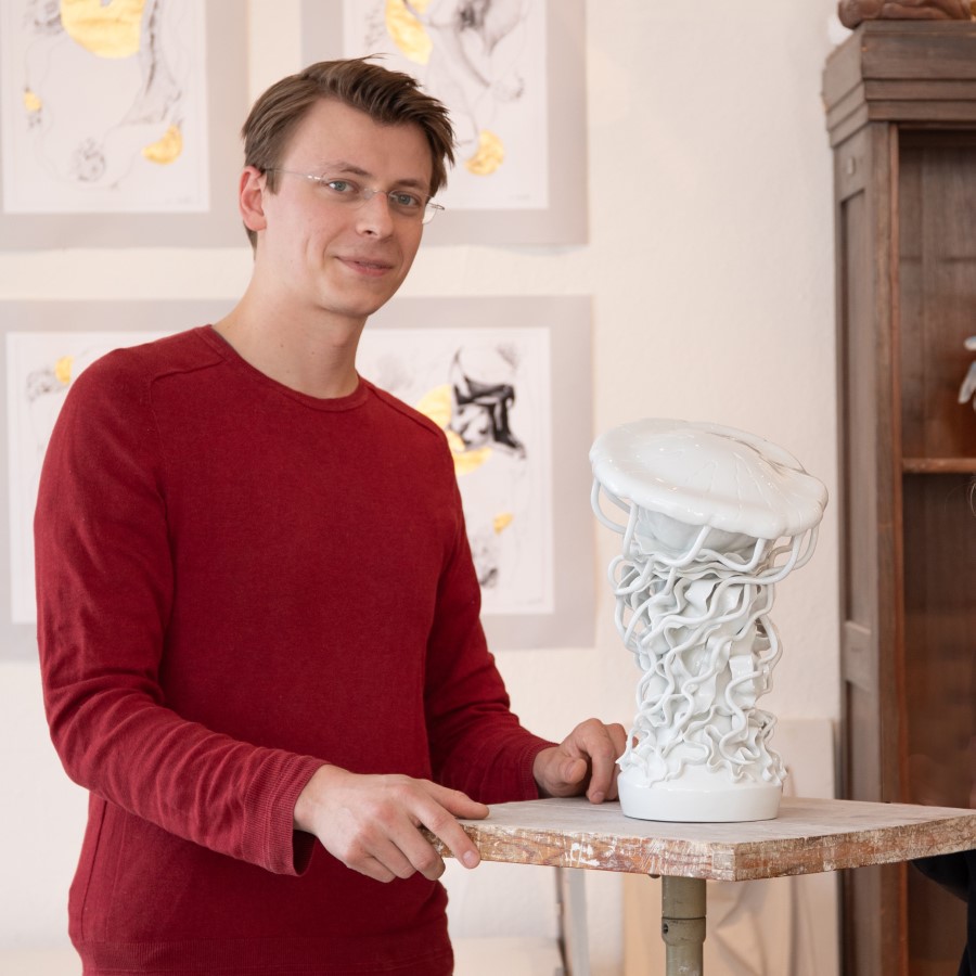Ein junger Mann steht vor einem Kunstwerk aus weißem Porzellan und lächelt in die Kamera.