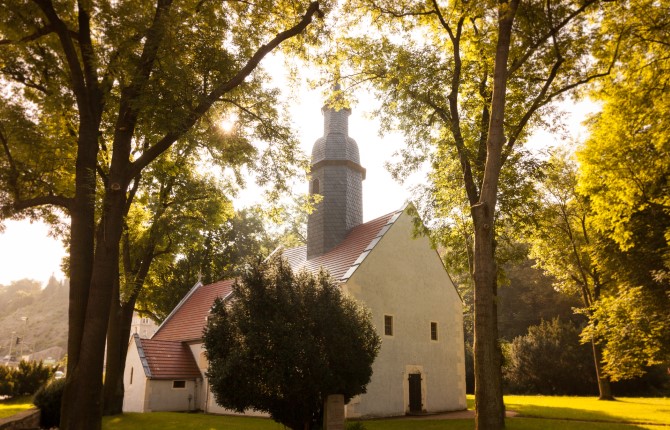Eine kleine weiße Kirche mit rotem Ziegeldach und schiefergedecktem Turm steht inmitten einer Parkanlage.