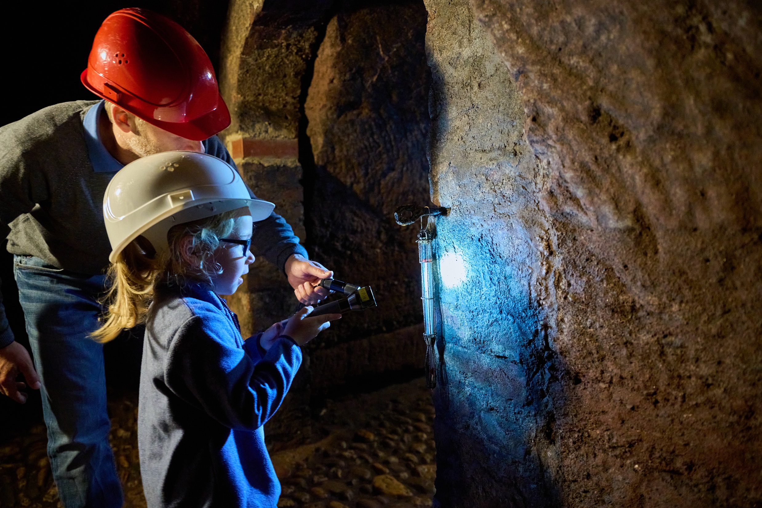 Ein Kind mit Helm leuchtet mit einer Taschenlampe Gestein unter dem Schloss an, hinter ihm ein Erwachsener mit Helm.