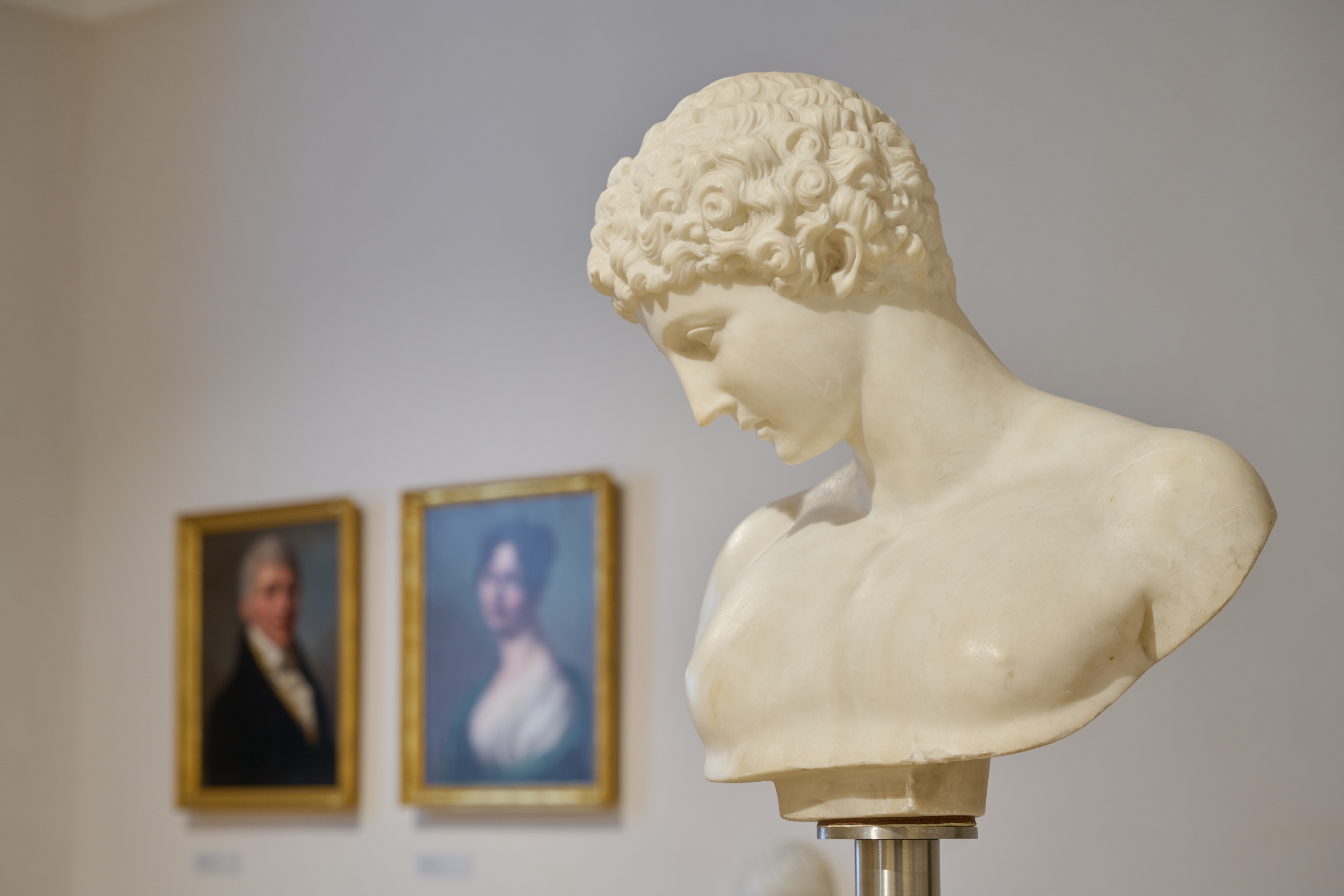 In einem Ausstellungsraum sind drei Exponate zu sehen: im Vordergrund die antik anmutende Büste eines nachdenklich schräg nach unten schauenden jungen Mannes und im Hintergrund zwei gerahmte Porträtmalereien.