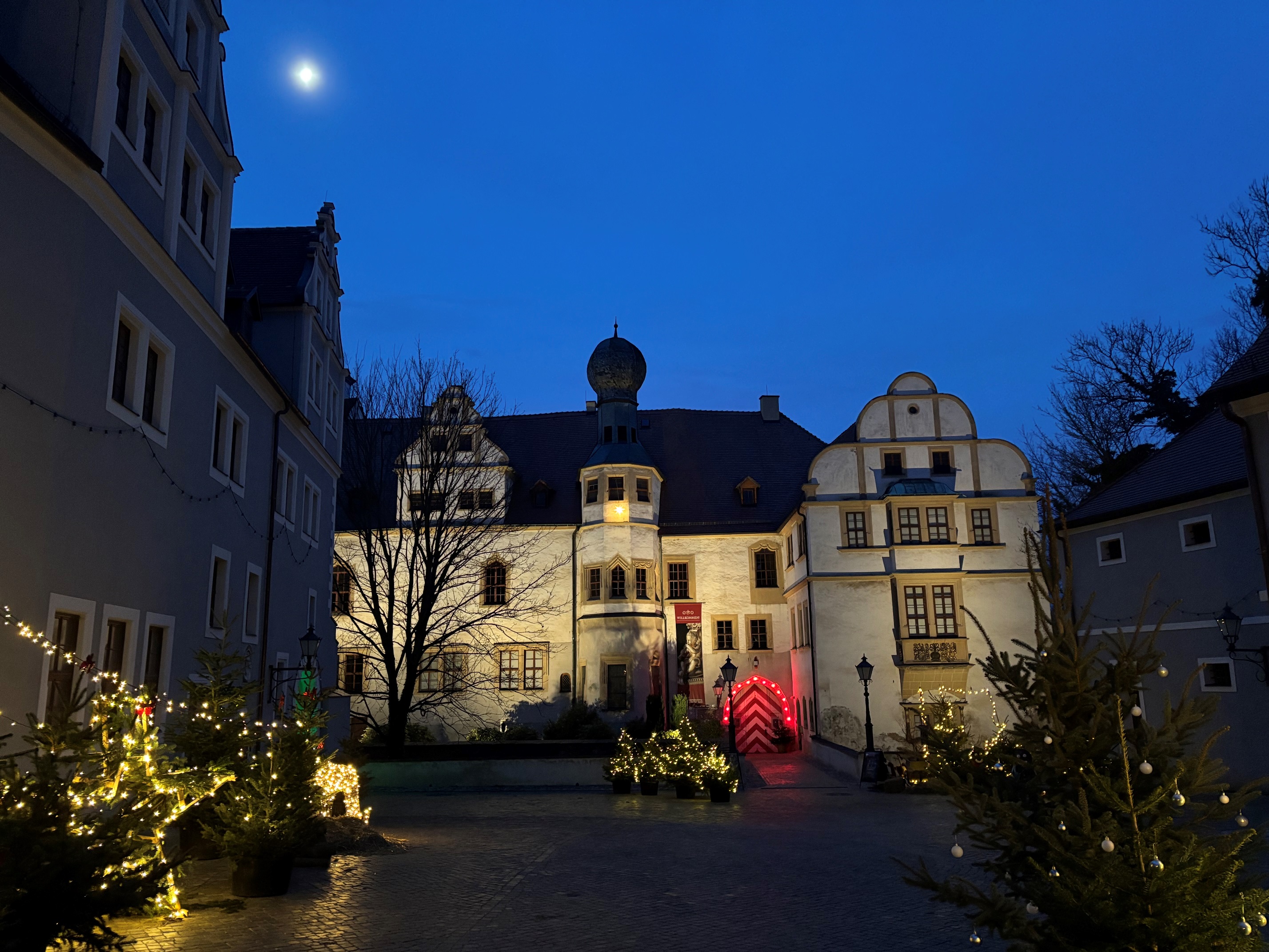 Spätabendliche Aufnahme eines historischen, weihnachtlich beleuchteten Schlosshofes. Am klaren, dunkelblauen Himmel scheint der Mond.
