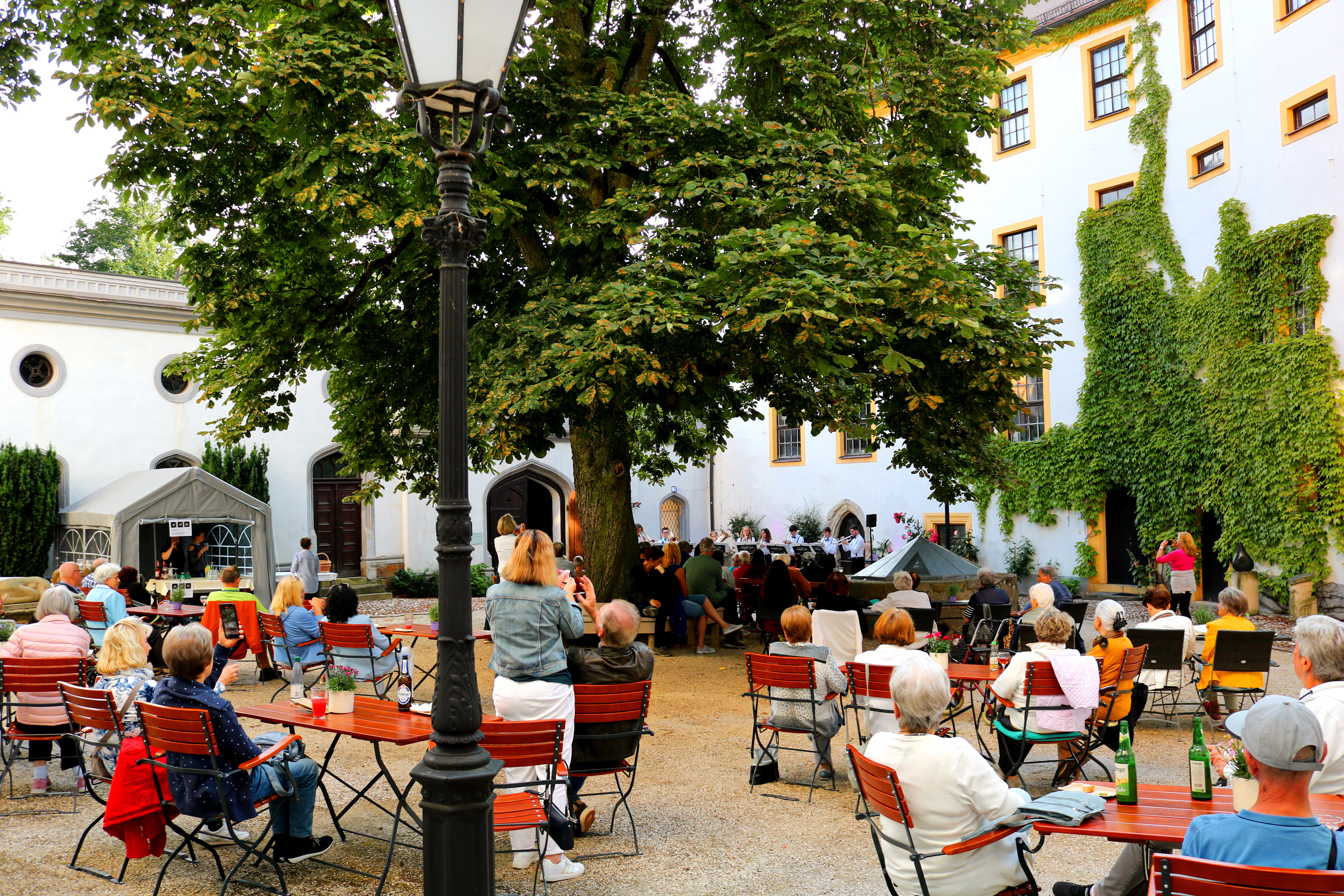 Viele Menschen sitzen in einem mit Café-Bestuhlung ausgestatteten Schlosshof. Unter einem großen Laubbaum geben Musiker ein Konzert.