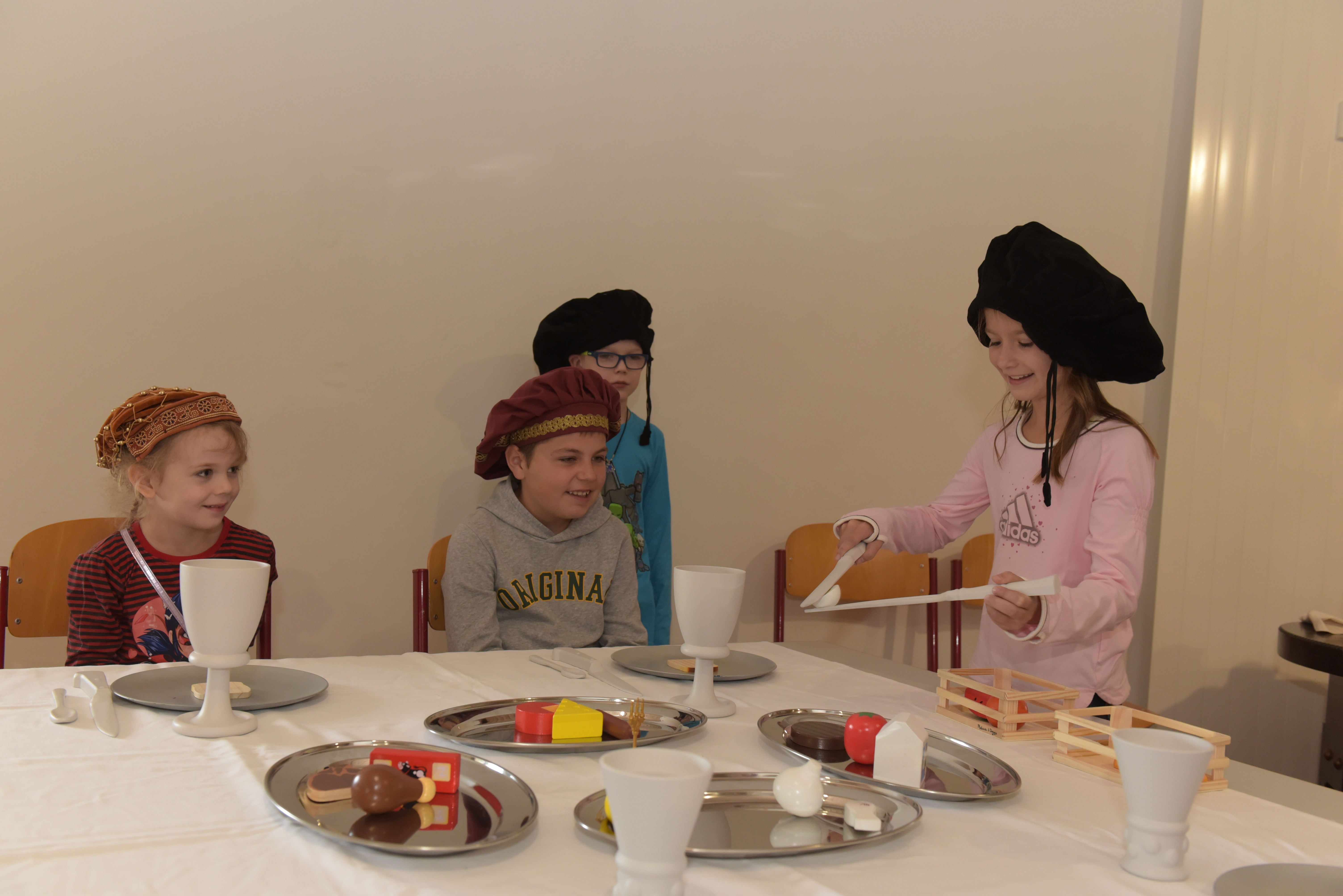 Vier Kinder mit Kopfbedeckungen im Renaissance-Stil üben sich an einem Tisch in historischer Esskultur
