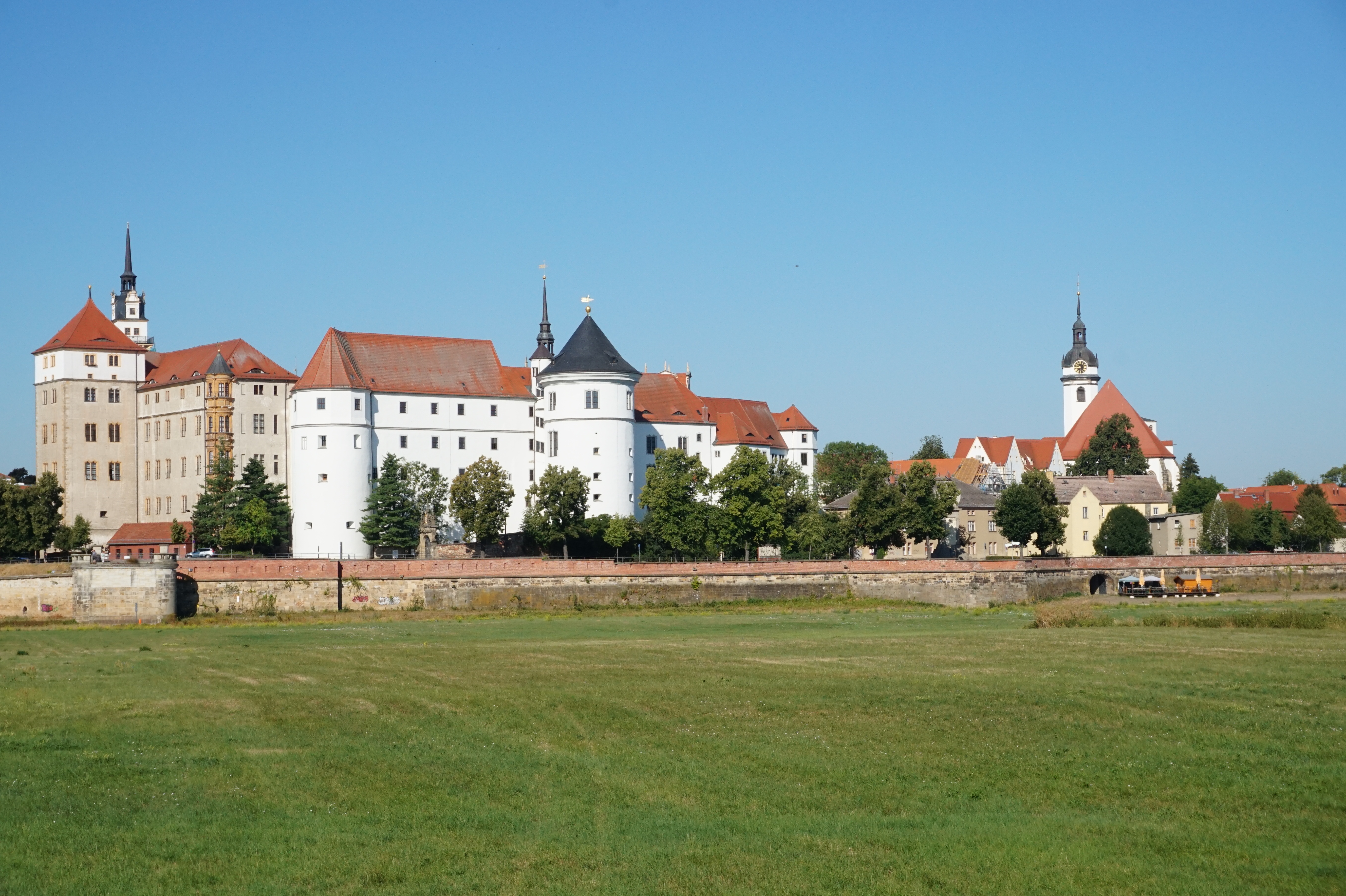 Sommerliche Außenansicht eines großen Schlosskomplexes mit weißer Fassade und mehreren Türmen, im Vordergrund eine flache Grasfläche