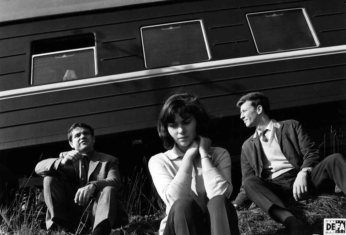 Auf dem Gras einen Bahndamms sitzen vor einem Eisenbahnwaggon eine junge Frau und zwei junge Männer.