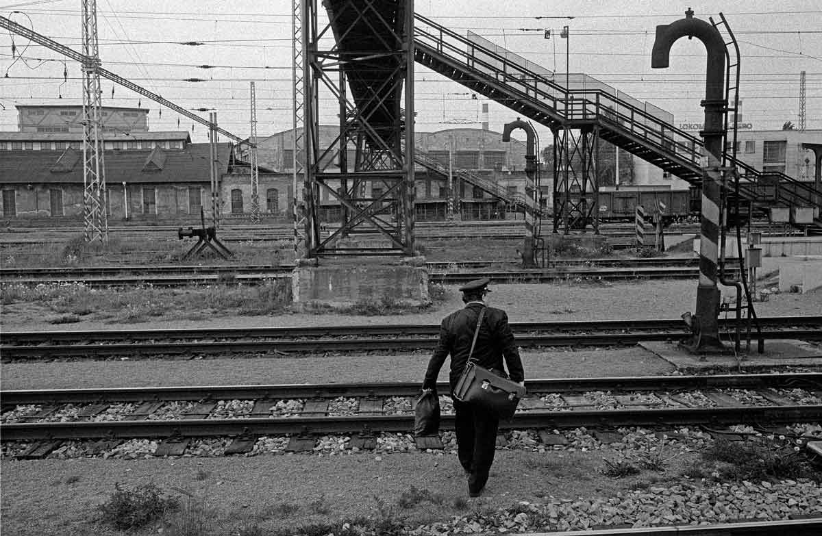 Schwarzweißfotografie eines Mannes, der die Bahngleise eines trostlos wirkenden Bahnhofsbereiches überquert