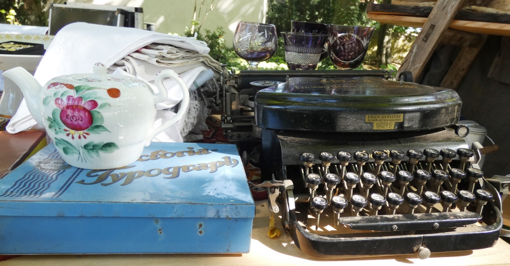Nahaufnahme eines Flohmarkt-Tisches, auf dem neben einer historischen Schreibmaschine ein mit Blumendekor verziertes Teekännchen, eine hellblauer Blechdose, ein kleiner Stapel weißer Tischdecken sowie einige geschliffene Weingläser stehen