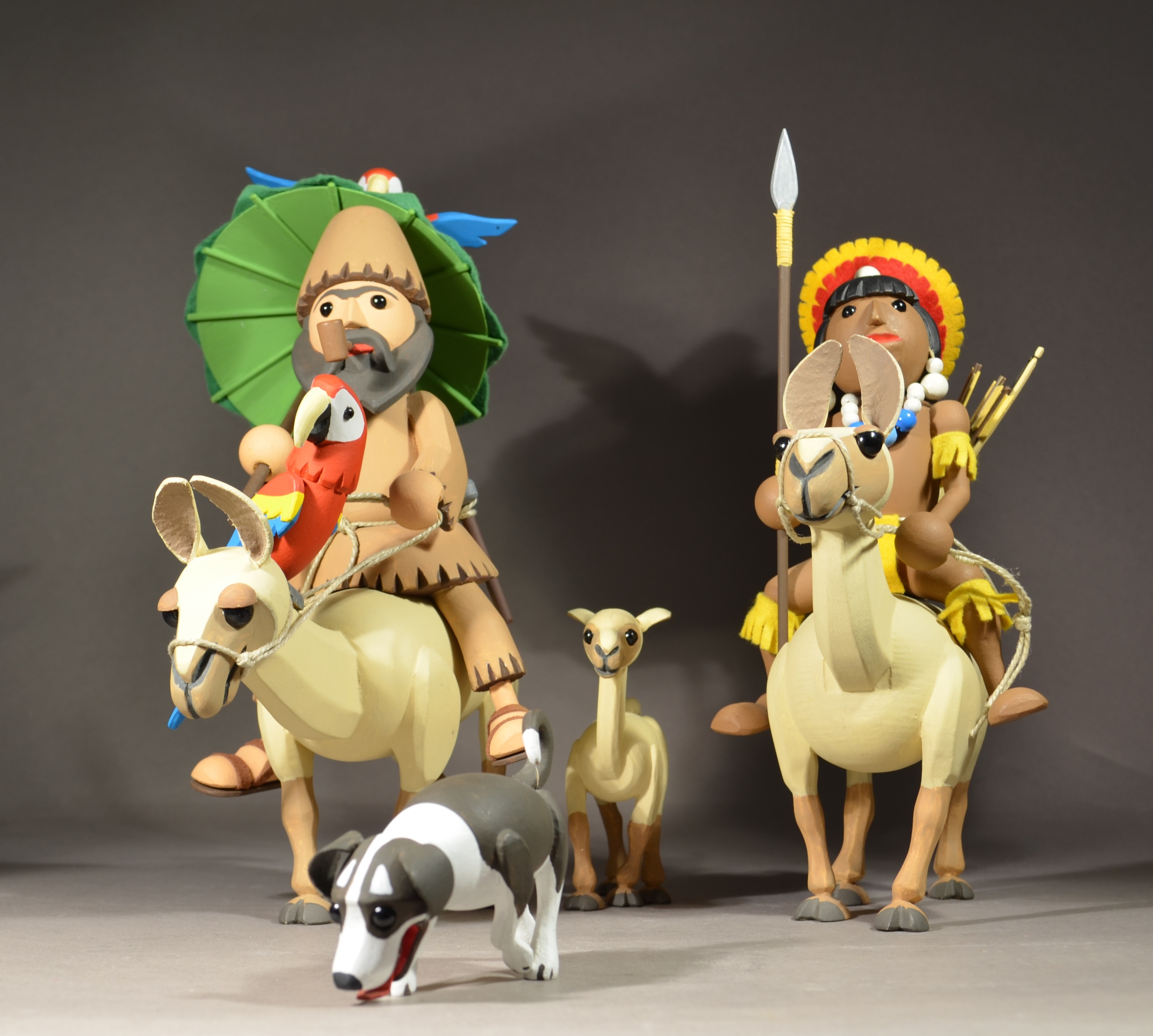 Zwei bunt bemalte männliche Figuren reiten auf Lamas: die bärtige Figur raucht dabei Pfeife und hat einen roten Papagei vor sich sitzen, die zweite Figur trägt indigen anmutende Kleidung. Zwischen den beiden laufen ein Hund und ein junges Lama.