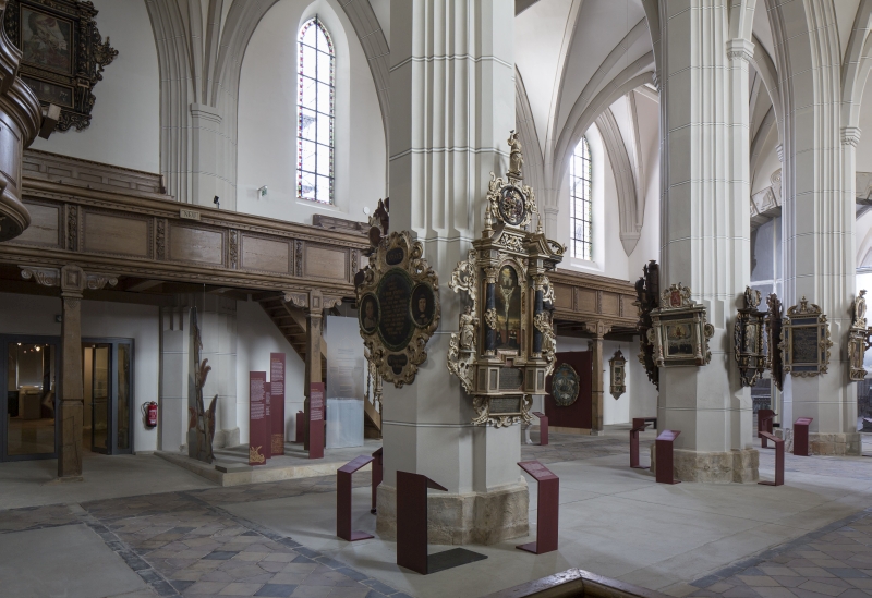 Ein hoch aufragendes Kirchenschiff mit weißen Wänden und einer hölzernen Empore. An den gotischen Säulen sind eine Vielzahl von Gemälden in reich verzierten Rahmen angebracht.