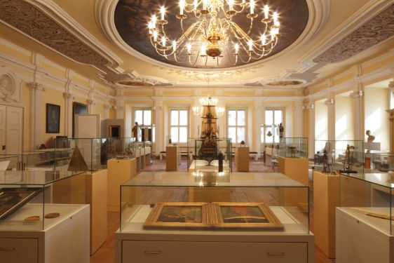 Foto eines prächtigen Barocksaals, in dem sich eine Vielzahl von Vitrinen mit unterschiedlichen Exponaten befinden und von dessen bemalter und reich verzierter Decke ein großer Kronleuchter hängt