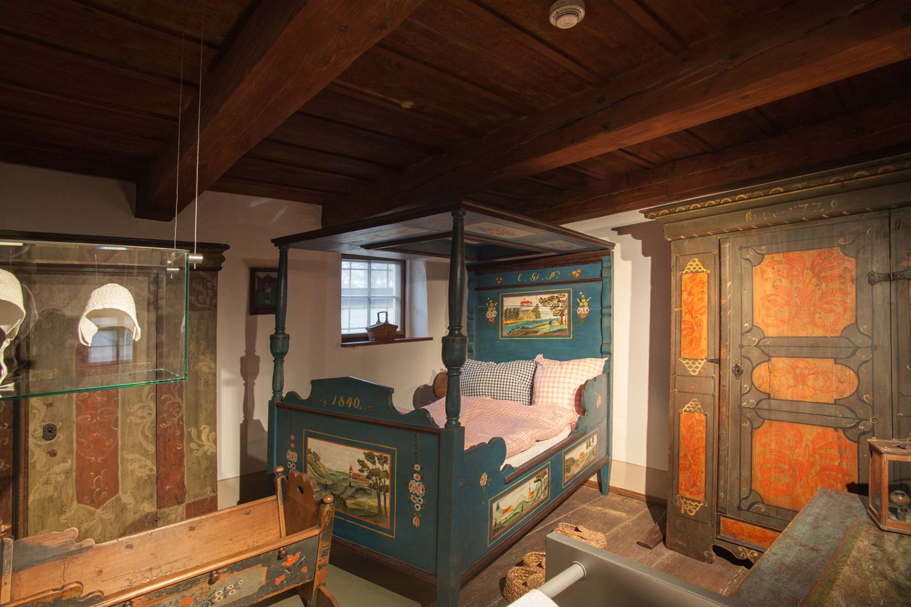 Inszenierung einer bäuerlich anmutenden Schlafstube mit handbemalten Holzmöbeln: ein Doppelbett mit Baldachin, eine Wiege und zwei Schränke stehen dicht beieinander. Der Raum ist niedrig, oben befindet sich eine Holzbalkendecke.