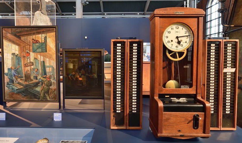 Eine historische Pendeluhr in einem Holzgehäuse steht in der Dauerausstellung neben dazugehörigem technischen Gerät; links daneben zwei Gemälde, die Industrieszenen zeigen.