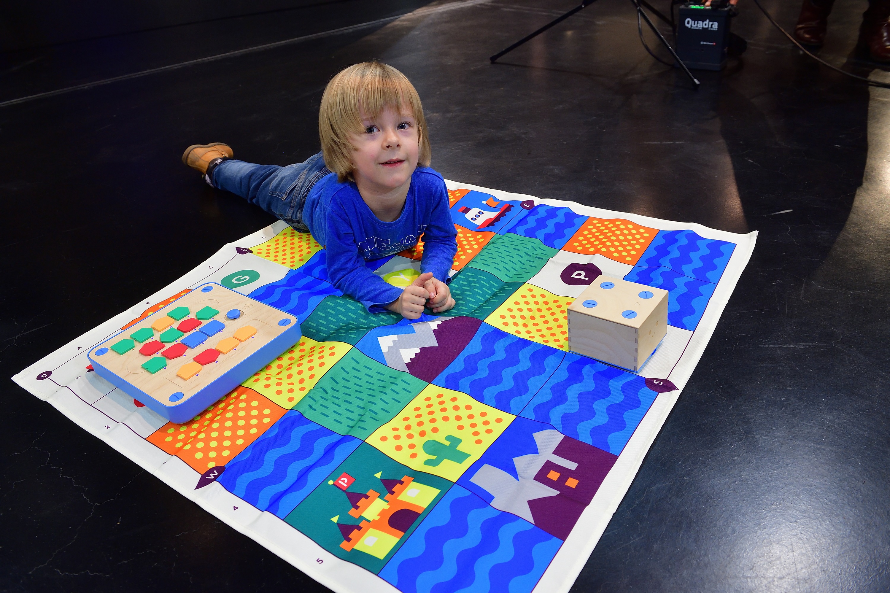 Ein kleines Kind liegt auf einem bunten Spielteppich und blickt verschmitzt in die Kamera.