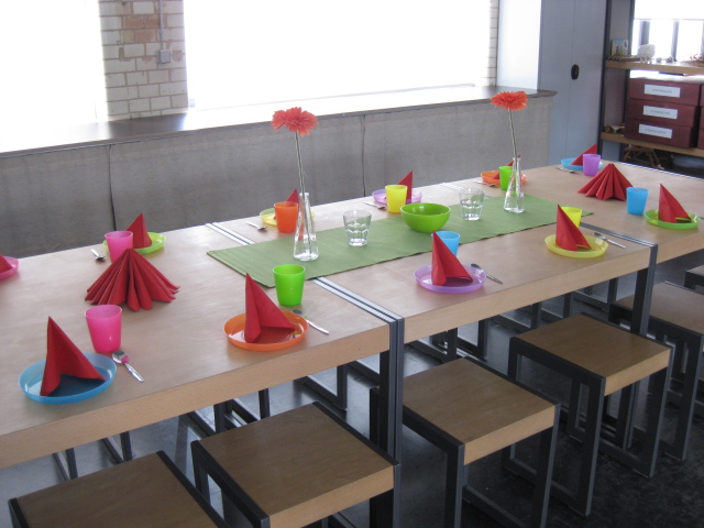 Ein langer Tisch ist mit bunten Plastiktellern und -bechern, roten Servietten und zwei kleinen Blumenvasen geschmückt, unter dem Tisch stehen Hocker.