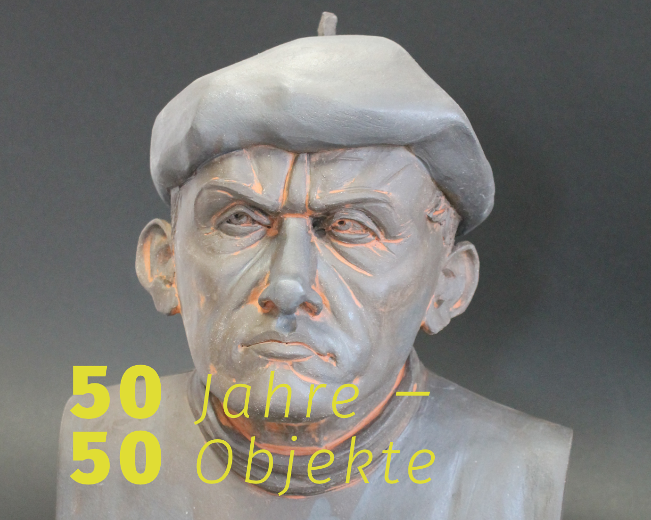 Auf dem Foto einer geschnitzten Büste eines Mannes mit Baskenmütze steht in Gelb der Schriftzug Jubiläumsausstellung „50 Jahre – 50 Objekte“.