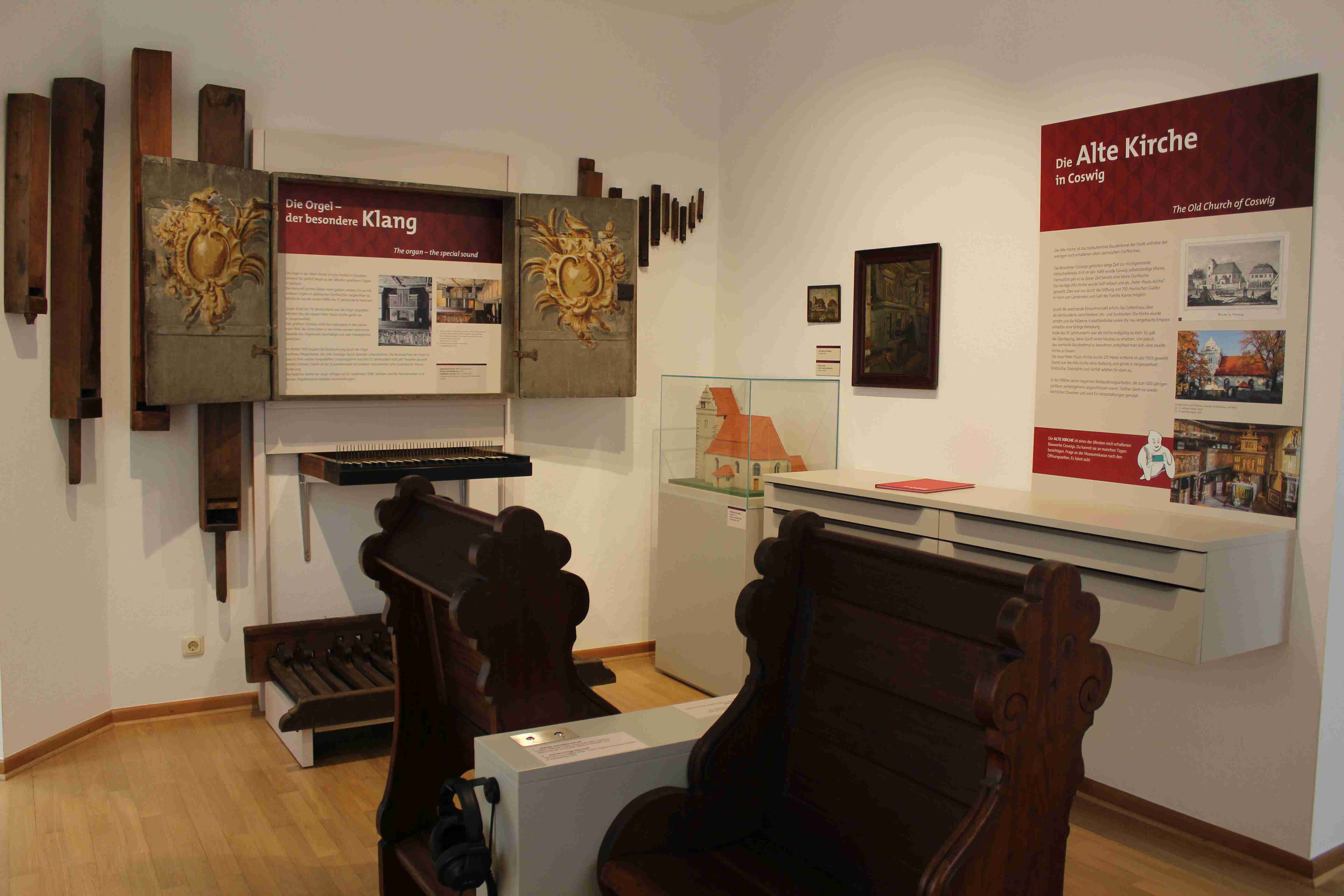 Das Foto zeigt neben historischem Kirchengestühl mehrere an der Wand angebrachte historische Orgelpfeifen, daneben Wandtexte, ein Schubladen-Element sowie eine Eck-Vitrine mit einem Modell der Coswiger Kirche.