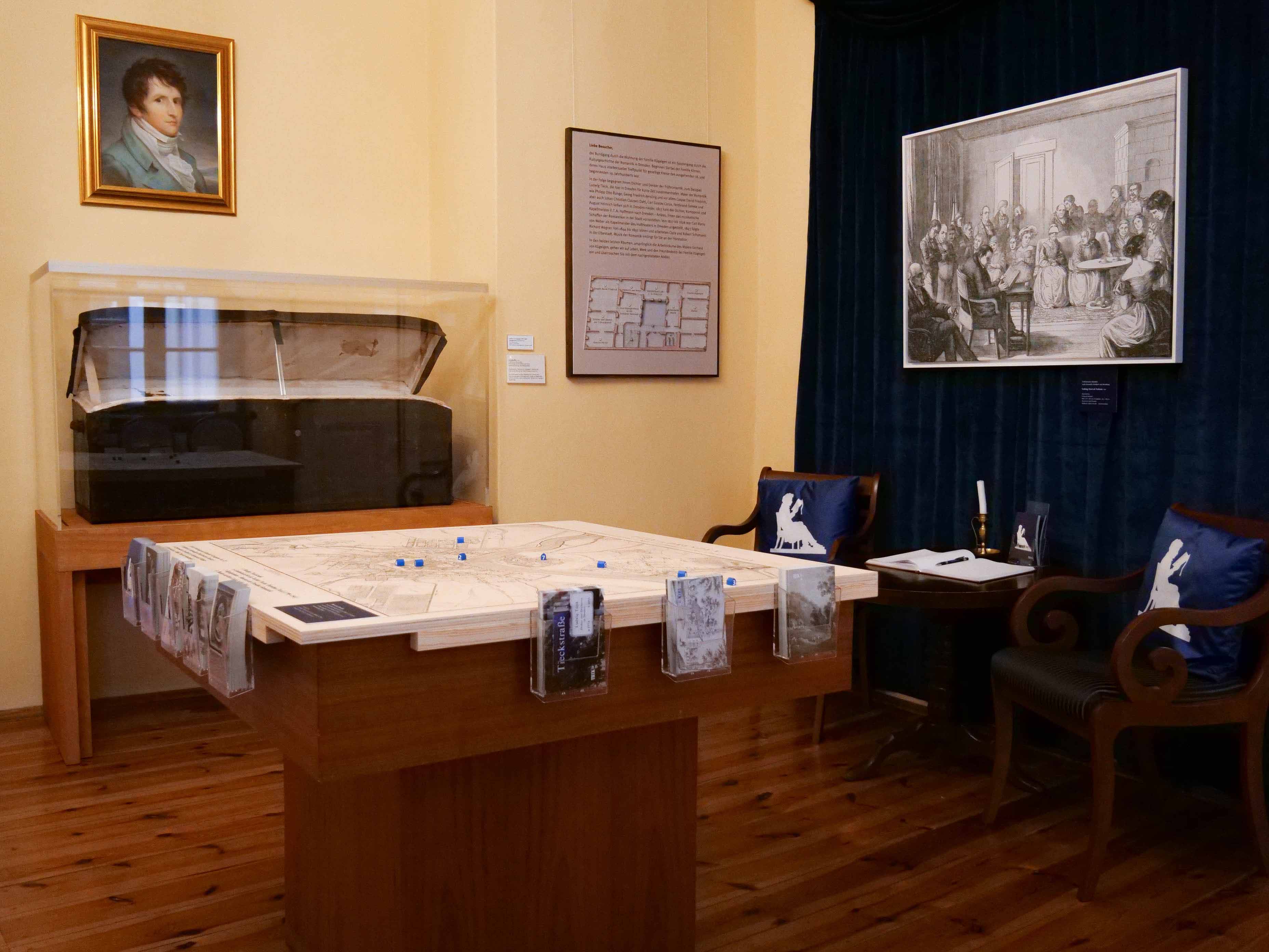 Foto eines Ausstellungsbereiches mit einem quadratischen Tisch in der Mitte, auf dem ein Stadtplan aufgebracht ist, an den Seiten sind Postkartenhalter. Im Hintergrund befindet sich eine Vitrine mit einer aufgeklappten historischen Truhe.
