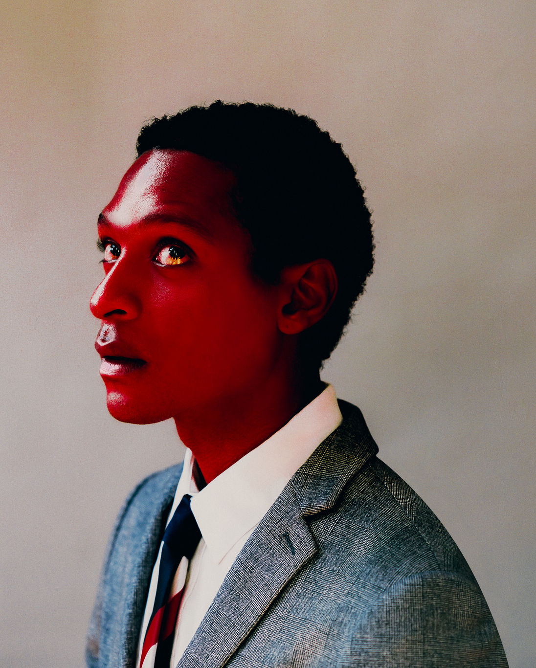 Diese Fotografie von Aaron Ricketts zeigt Kopf- und Schulterpartie eines jungen afrikanischen oder afroamerikanischen Mannes im Seitenprofil. Seine Haut ist glänzend-rot gefärbt. Gekleidet in Anzug und Krawatte, richtet er seinen Blick nach schräg oben.