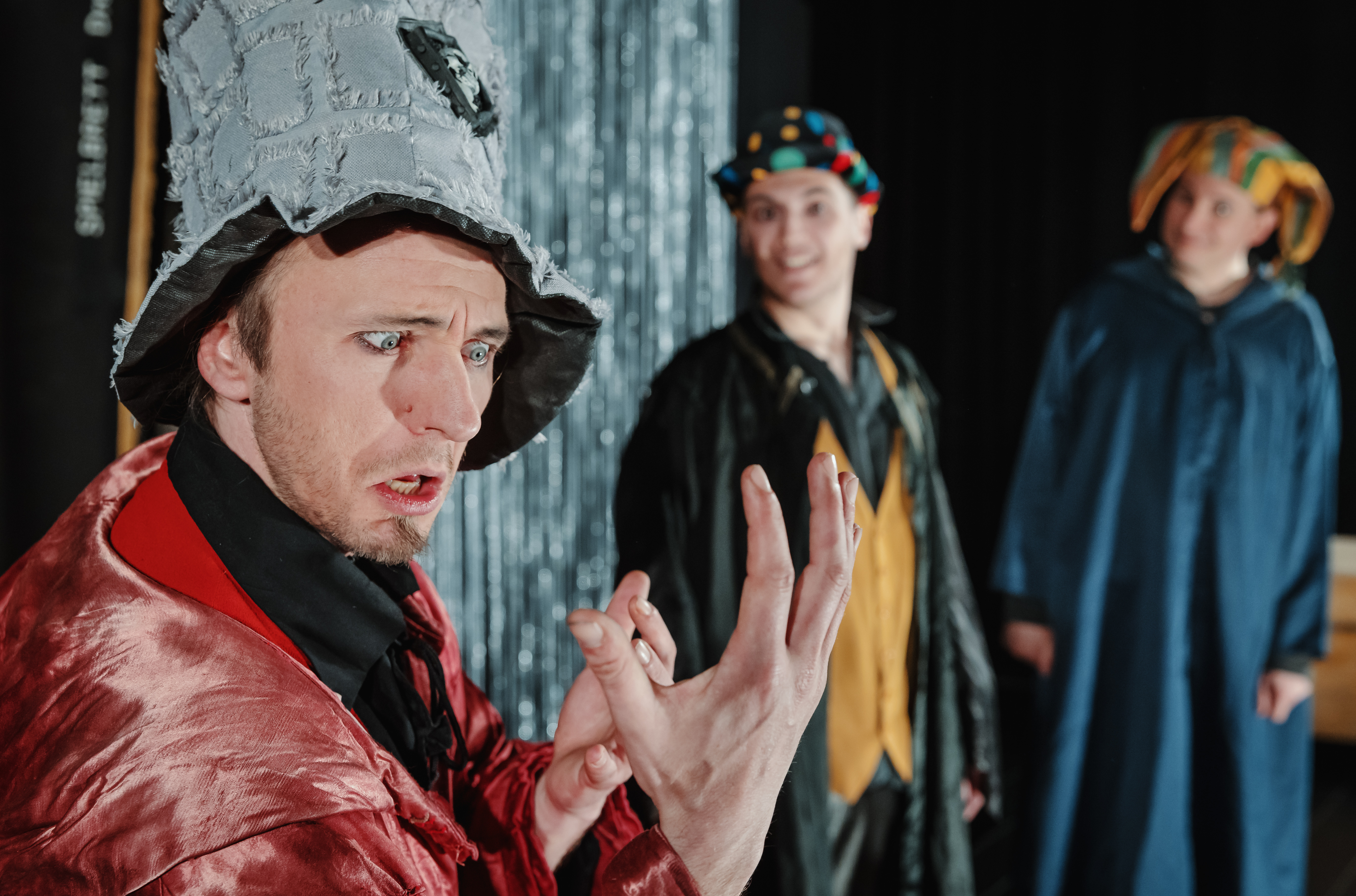 Ein in rote Seidenrobe und grauen Stoffzylinder gekleideter Schauspieler schaut ausdrucksstark in seine geöffnete Hand, im Hintergrund sind zwei weitere Schauspieler zu erkennen, die ihn anblicken.