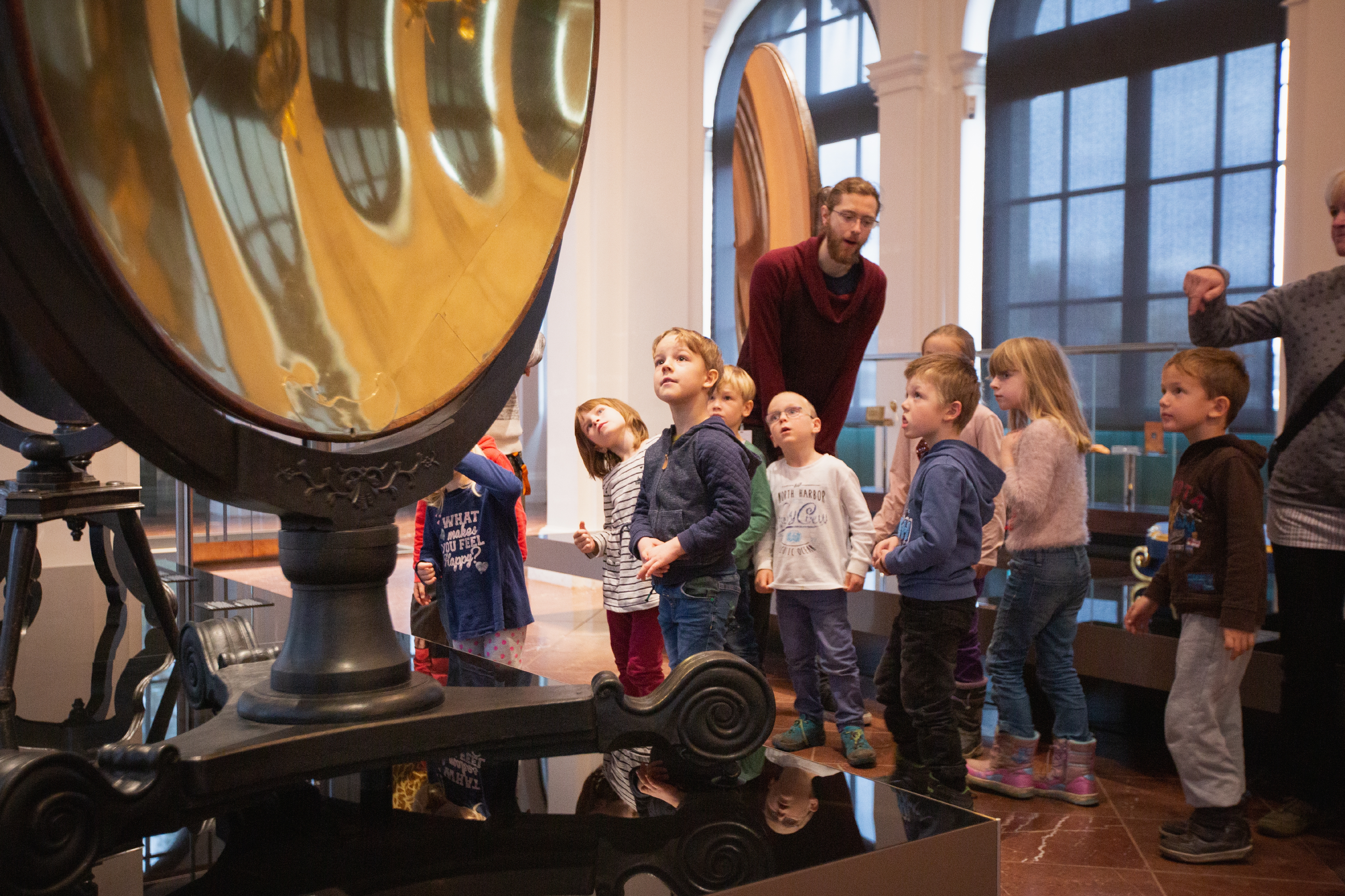 Das Foto zeigt eine Gruppe von Vorschulkindern mit einem Museumspädagogen bei einem Vermittlungsangebot im Mathematisch-Physikalischen Salon. Die Kinder bestaunen einen riesigen historischen Hohlspiegel.