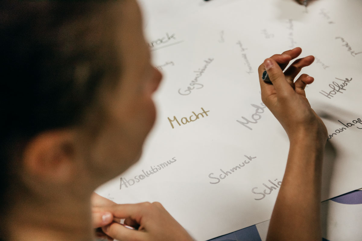 Das Foto zeigt die einen Stift haltende Hand eines Mädchens über einem weißen Papierbogen, auf dem in silberner und goldener Farbe Worte wie „Mode“, „Macht“, „Schmuck“ und „Absolutismus“ geschrieben wurden.
