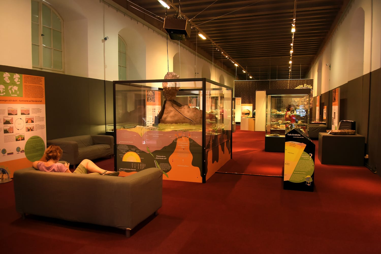 Blick in die Ausstellung „Bewegte Erde – Eine geologische Zeitreise“ mit mehreren Vitrinen und Tafeln und einer Medienstation; im Vordergrund ist eine auf einem Sofa sitzende Besucherin zu sehen.